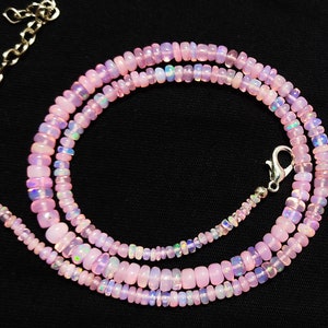 May include: A delicate necklace composed of small, light pink opal beads. The beads are strung together, creating a layered effect. The necklace has a silver-colored chain and clasp, and is displayed against a black background.