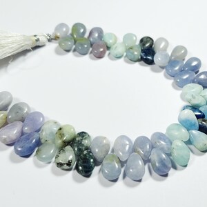 May include: A strand of teardrop-shaped gemstones in shades of blue, green, and purple, with a white tassel at one end. The stones vary in color, creating a gradient effect. The gemstones are arranged in a curved line.