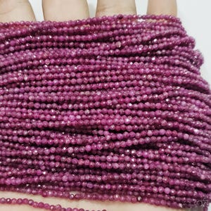 Puede incluir: Primer plano de una colección de pequeñas cuentas facetadas de color rubí. Las cuentas están ensartadas juntas, formando múltiples hebras. Un borla roja es visible en el lado izquierdo. Las cuentas son de un color rosa intenso y rico.