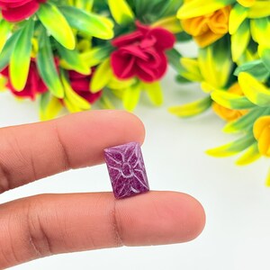 May include: A small, rectangular, deep magenta gemstone featuring a carved floral pattern on its surface. The stone has a faceted texture and is held against a background of vibrant red and yellow artificial flowers with green leaves.