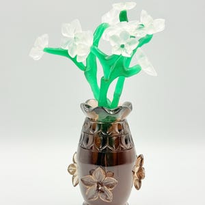 Puede incluir: Un jarrón decorativo de cristal marrón oscuro, adornado con tres detalles florales. Contiene un ramo de flores de cristal blanco con tallos verdes, creando un contraste llamativo con el color del jarrón.