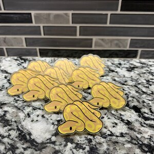 May include: A set of eight yellow and brown cartoon snake stickers. The stickers are glossy and have a clear background.