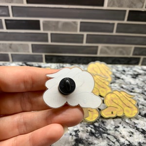 May include: A set of four yellow and white snake enamel pins. The pins are in the shape of a snake curled up. The back of one pin is shown with a black pin back.