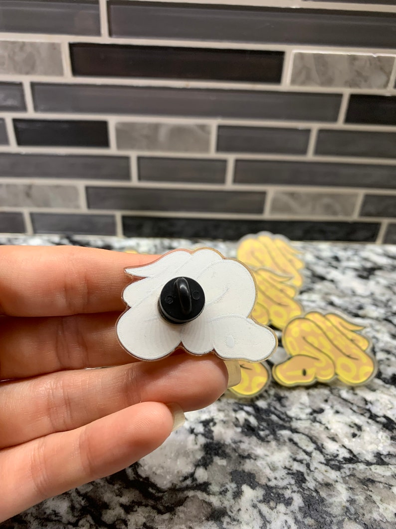 May include: A set of four yellow and white snake enamel pins. The pins are in the shape of a snake curled up. The back of one pin is shown with a black pin back.