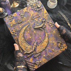 Può includere: Un diario fatto a mano con un tema celeste. La copertina presenta un design strutturato con una luna crescente, una balena e stelle in tonalità oro e viola. Accenti e abbellimenti metallici decorativi aggiungono all'estetica mistica.