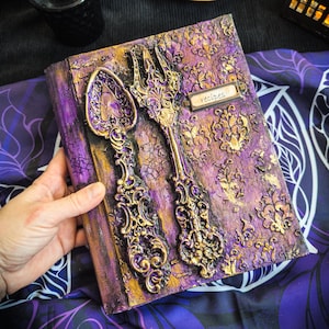 Peut inclure: Un livre de recettes violet et doré avec des ornements de cuillère et de fourchette. Le livre a une finition texturée et craquelée et une petite étiquette métallique qui dit "recipes". L'arrière-plan comprend une bouteille noire, un plat doré avec des bougies et un tissu violet à motifs.
