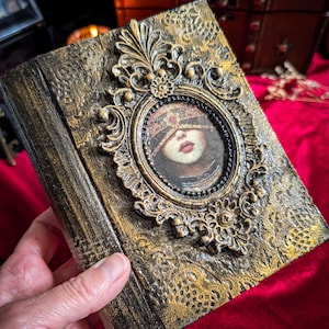 Peut inclure: Un livre rectangulaire avec une couverture texturée noire et dorée ornée d'un cadre ovale en relief. À l'intérieur du cadre se trouve l'image d'une personne portant un bandeau sur les yeux. La couverture présente des motifs de dentelle complexes.