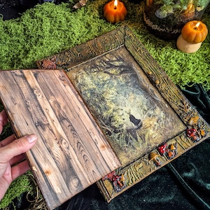 May include: A decorative wooden book with a forest scene illustration. The frame is adorned with mushrooms and leaves. The open book reveals a wood-grain pattern on the left page. Two pumpkin-shaped candles and a terrarium are in the background.