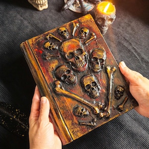 May include: A detailed, textured book cover with a skull and crossbones design. The cover is painted in shades of brown, orange, and red, with several small skulls and bone accents. The book is held by hands, suggesting a gothic or Halloween theme.
