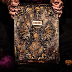 May include: A detailed, antique-style book with a textured, bronze-colored cover. The cover features embossed bird-like skeletal figures and the text "YOUR TEXT" on a small plaque. The book is held by hands with black nail polish and henna tattoos.