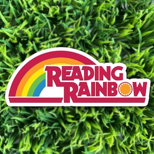 May include: A sticker with the words "Reading Rainbow" in red letters, with a rainbow graphic. The sticker has a white border and a sun graphic. The background is green.