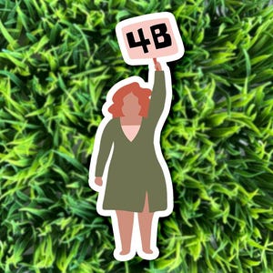 May include: A woman with red hair is holding a sign that says "4B" in black letters on a pink background. The woman is wearing a green jacket and is standing in front of a green background.