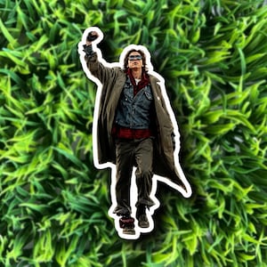 May include: A sticker featuring a person in a long coat, denim jacket, and sunglasses, with a raised fist. The person is wearing a red plaid shirt and dark pants. The sticker is set against a green background.