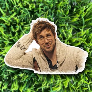 May include: A die-cut sticker featuring a man with light brown hair and glasses, wearing a cream-colored sweater. The sticker is placed on a background of green artificial grass.