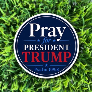 May include: A round sticker with a navy blue border and background. The text "Pray for PRESIDENT TRUMP" is in white and red. The bottom of the sticker reads "Psalm 109:8". The sticker is set against a background of green grass.