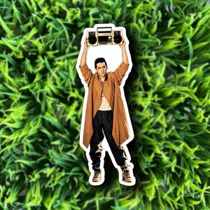 May include: A sticker featuring a person holding a boombox above their head. The person is wearing a brown coat, black pants, and tan boots. The boombox is beige and black. The sticker is on a green background.