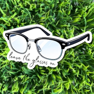 May include: A sticker featuring a pair of black-rimmed glasses with clear lenses. The text "leave the glasses on" is written in cursive below the glasses. The sticker is set against a background of green foliage.