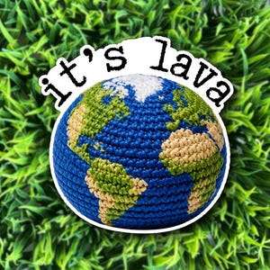 May include: A crocheted Earth globe with blue oceans, green and tan continents, and a white border. The text "it's lava" is above the globe. The background is green.