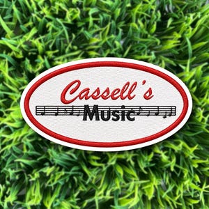 May include: An oval-shaped embroidered patch with a white background and a red border. The text 'Cassell's' is stitched in red script above the word 'Music' in black, which is centered over a black musical staff with notes.