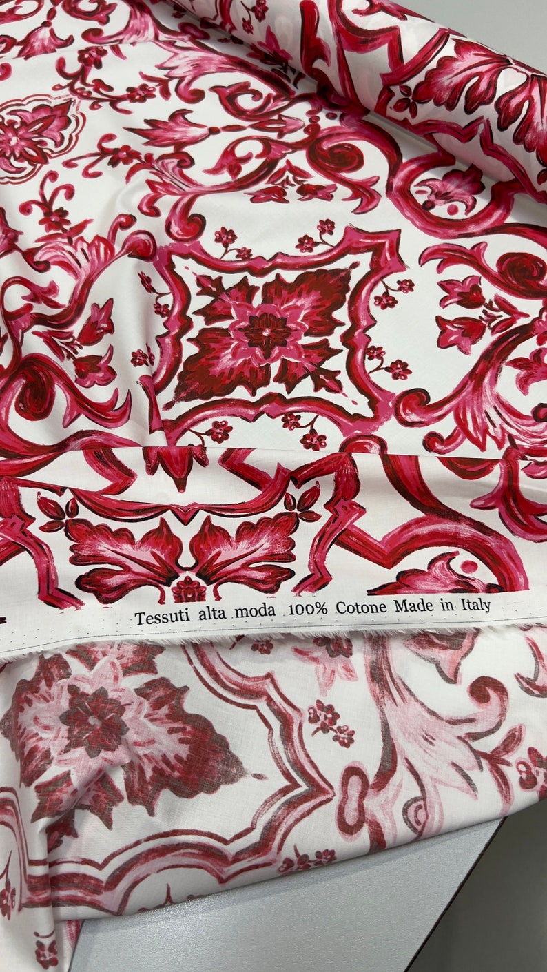 May include: White fabric with a red and pink floral pattern. The fabric has a label that reads "Tessuti alta moda 100% Cotone Made in Italy".