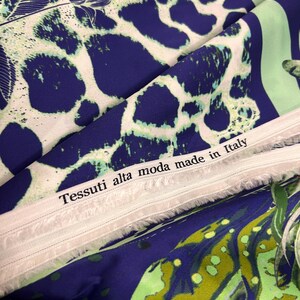 Op de afbeelding: Close-up van een stof met een patroon. Het ontwerp heeft grote, onregelmatige blauwe vormen tegen een witte achtergrond, met groene accenten. De tekst "Tessuti alta moda made in Italy" is zichtbaar.