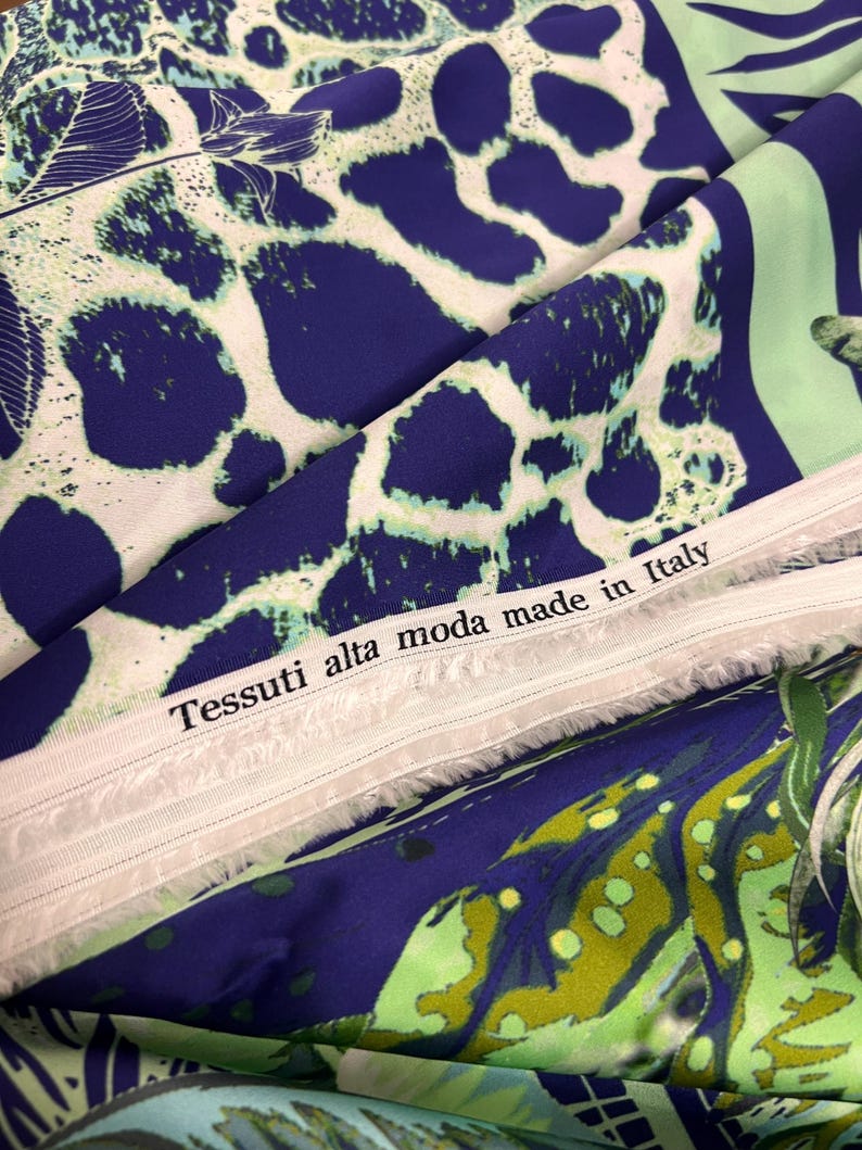 Op de afbeelding: Close-up van een stof met een patroon. Het ontwerp heeft grote, onregelmatige blauwe vormen tegen een witte achtergrond, met groene accenten. De tekst "Tessuti alta moda made in Italy" is zichtbaar.