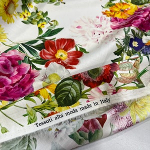 May include: White fabric with a floral print featuring various flowers in shades of pink, red, yellow, and blue. The fabric has a label that reads "Tessuti alta moda made in Italy."