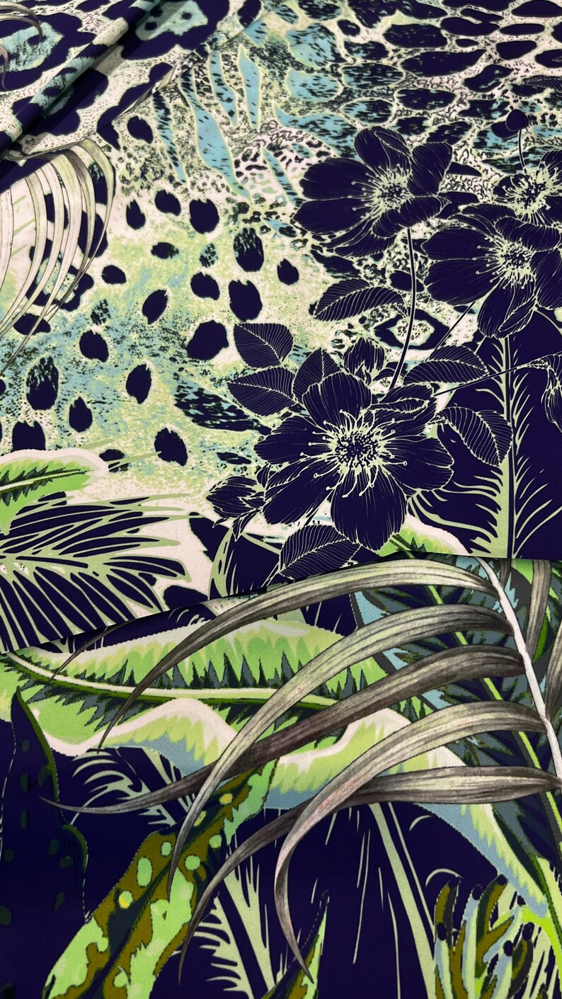 Op de afbeelding: Een stof met een donkerblauwe achtergrond met een bloemen- en bladerpatroon in wit en groen. Het ontwerp omvat grote, gestileerde bloemen en tropisch gebladerte, met een luipaardprint-achtig patroon op de achtergrond.