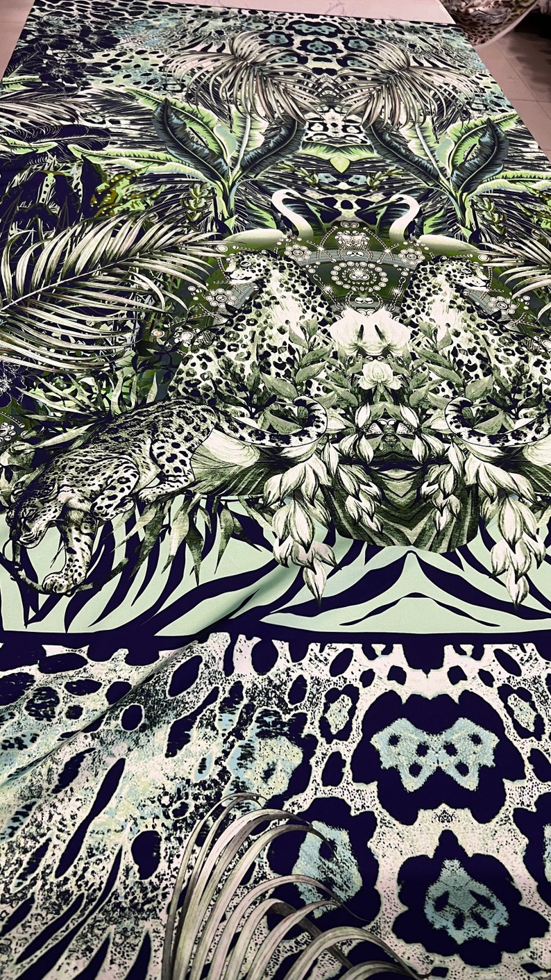Op de afbeelding: Stof met een jungle-thema print. Het ontwerp toont een luipaard, tropische bladeren en zebra strepen in tinten groen, wit en marineblauw. Het patroon herhaalt zich over de stof, waardoor een visueel rijk en gedetailleerd textiel ontstaat.