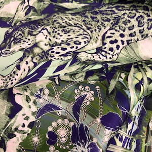 Op de afbeelding: Een stof met een jungle-thema. Het ontwerp toont een luipaard te midden van groene en blauwe bladeren, met bloemen- en kettingpatronen. Het gedetailleerde ontwerp van de stof cre&euml;ert een levendig textiel.