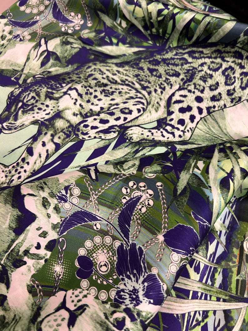 Op de afbeelding: Een stof met een jungle-thema. Het ontwerp toont een luipaard te midden van groene en blauwe bladeren, met bloemen- en kettingpatronen. Het gedetailleerde ontwerp van de stof cre&euml;ert een levendig textiel.