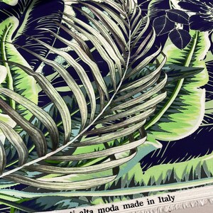 Op de afbeelding: Stof met een tropisch bladerenpatroon in groen, blauw en wit. De stof heeft grote palmbladeren en ander gebladerte tegen een donkerblauwe achtergrond. De tekst "Tessuti alta moda made in Italy" staat op de rand.