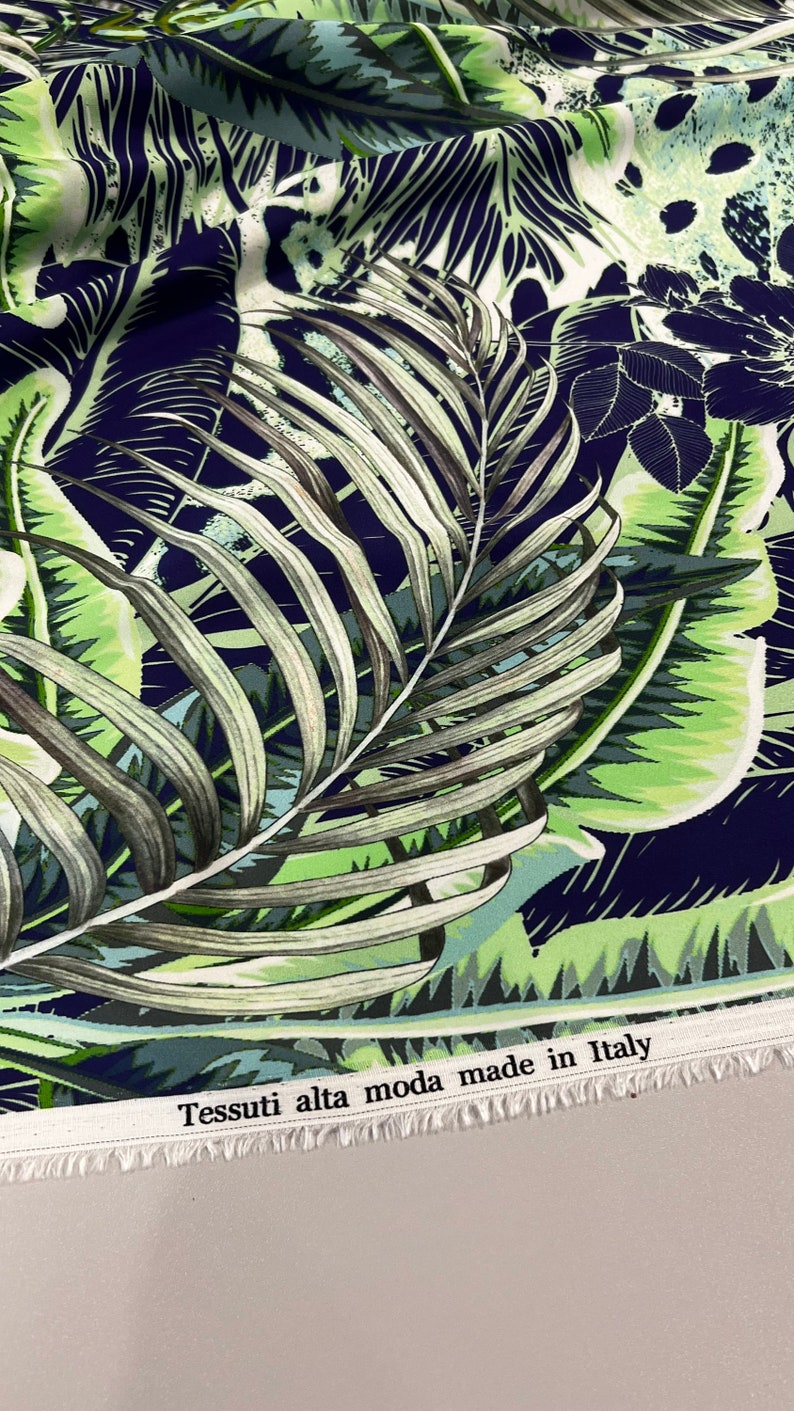 Op de afbeelding: Stof met een tropisch bladerenpatroon in groen, blauw en wit. De stof heeft grote palmbladeren en ander gebladerte tegen een donkerblauwe achtergrond. De tekst "Tessuti alta moda made in Italy" staat op de rand.