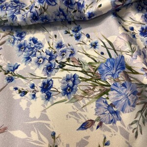 May include: A white fabric with a blue floral pattern. The flowers are a light blue and have a watercolor effect. The fabric has a subtle sheen.