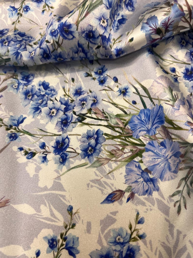 May include: A white fabric with a blue floral pattern. The flowers are a light blue and have a watercolor effect. The fabric has a subtle sheen.