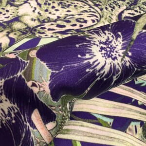 Op de afbeelding: Close-up van een stof met een levendige bloemen- en dierenprint. Het ontwerp toont grote paarse bloemen met witte centra en een luipaardprint in groen- en beigetinten. De stof heeft een zijdeachtige glans.