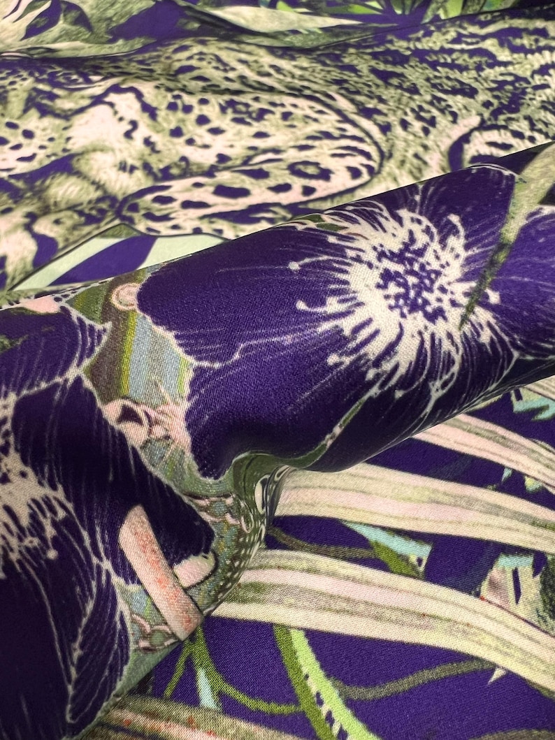 Op de afbeelding: Close-up van een stof met een levendige bloemen- en dierenprint. Het ontwerp toont grote paarse bloemen met witte centra en een luipaardprint in groen- en beigetinten. De stof heeft een zijdeachtige glans.