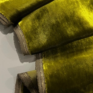 May include: Close-up of a folded piece of fabric in a bright, vibrant green colour. The fabric has a soft, velvety texture and a subtle sheen. The edges of the fabric are trimmed with a thin, gold-coloured thread.