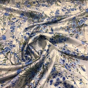 May include: A close-up of a white satin fabric with a blue floral print. The fabric is folded and draped, showing the texture and pattern.