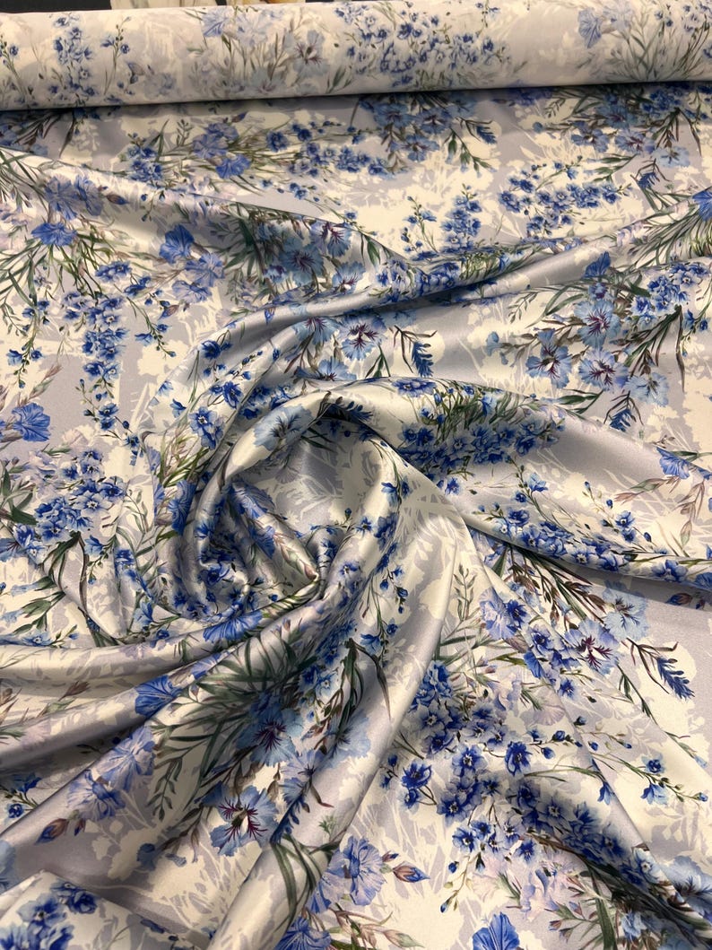 May include: A close-up of a white satin fabric with a blue floral print. The fabric is folded and draped, showing the texture and pattern.