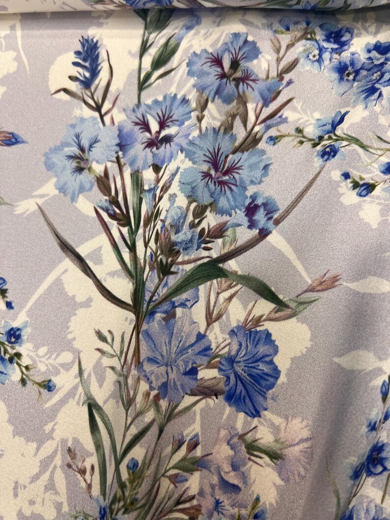 May include: A close-up of a light purple fabric with a floral print. The flowers are blue and white with green stems and leaves.
