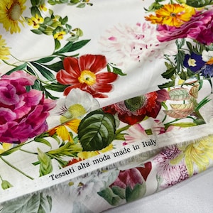 May include: White fabric with a floral print featuring various flowers in shades of pink, red, yellow, and blue. The fabric has a label that reads "Tessuti alta moda made in Italy."
