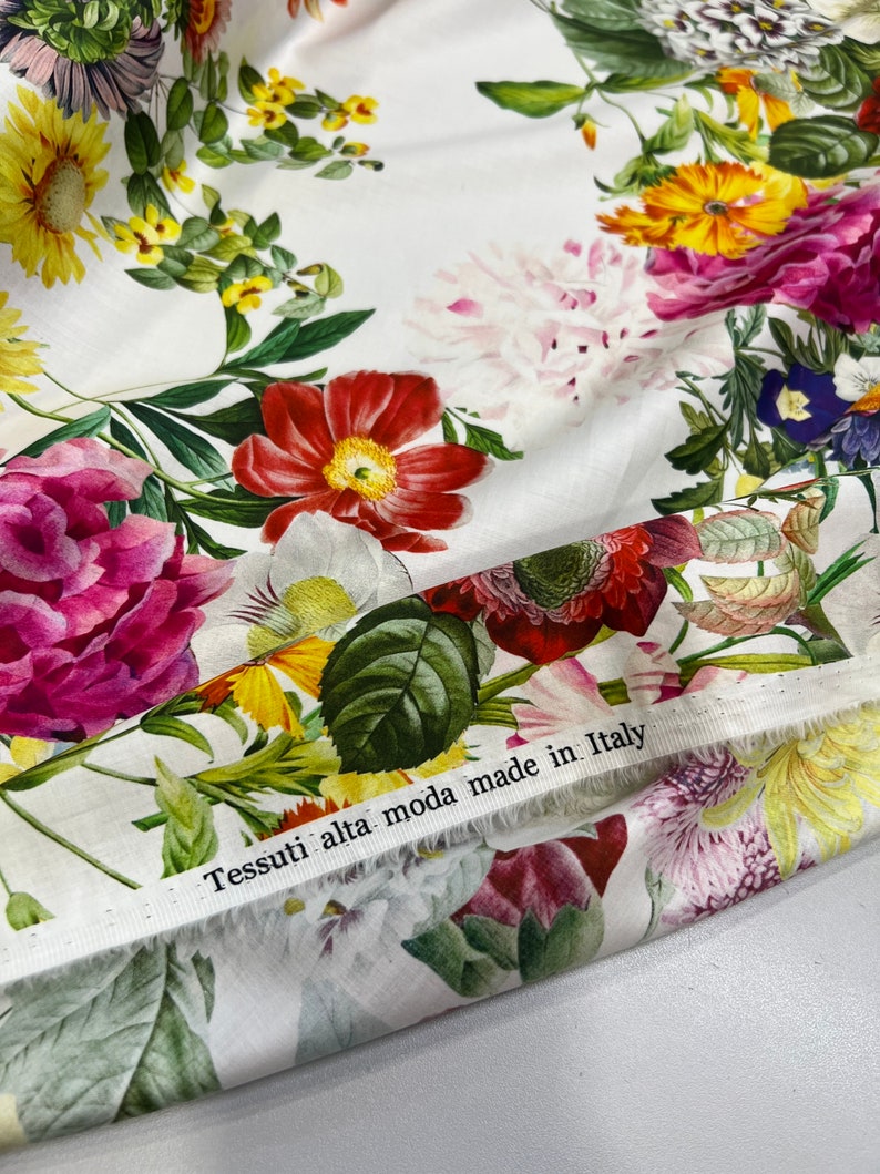 May include: White fabric with a floral print featuring various flowers in shades of pink, red, yellow, and blue. The fabric has a label that reads "Tessuti alta moda made in Italy."