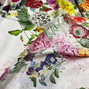 May include: White fabric with a floral print featuring a variety of flowers in shades of pink, red, yellow, and blue. The fabric is soft and has a linen-like texture.