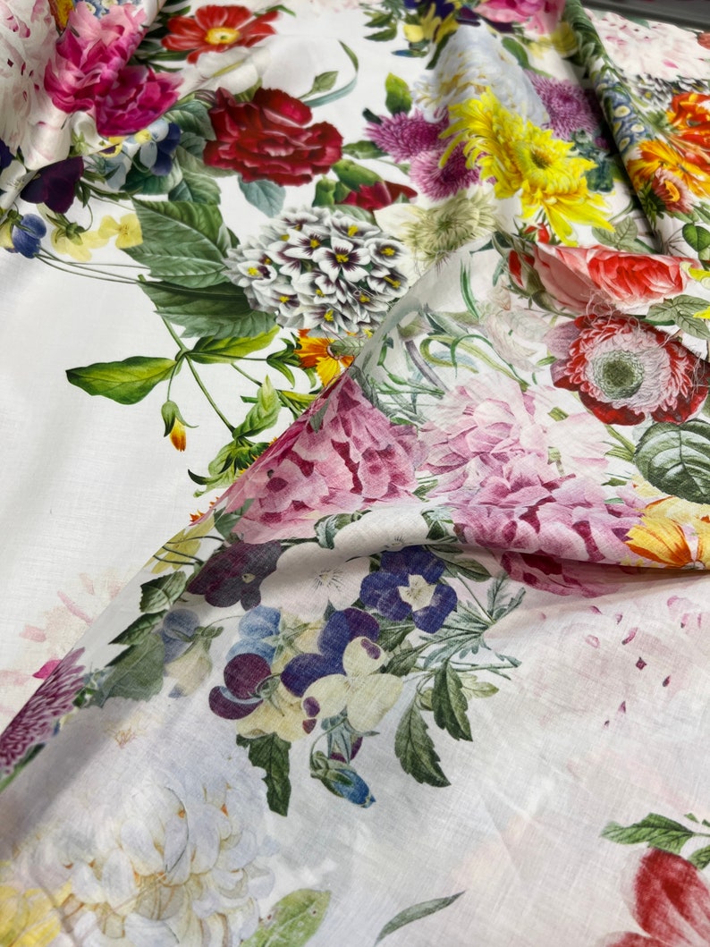 May include: White fabric with a floral print featuring a variety of flowers in shades of pink, red, yellow, and blue. The fabric is soft and has a linen-like texture.