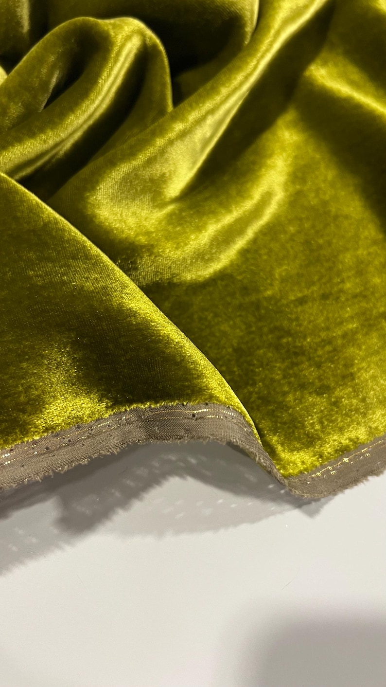 May include: A close-up of a bright green velvet fabric with a subtle sheen. The fabric has a decorative trim along the edge.