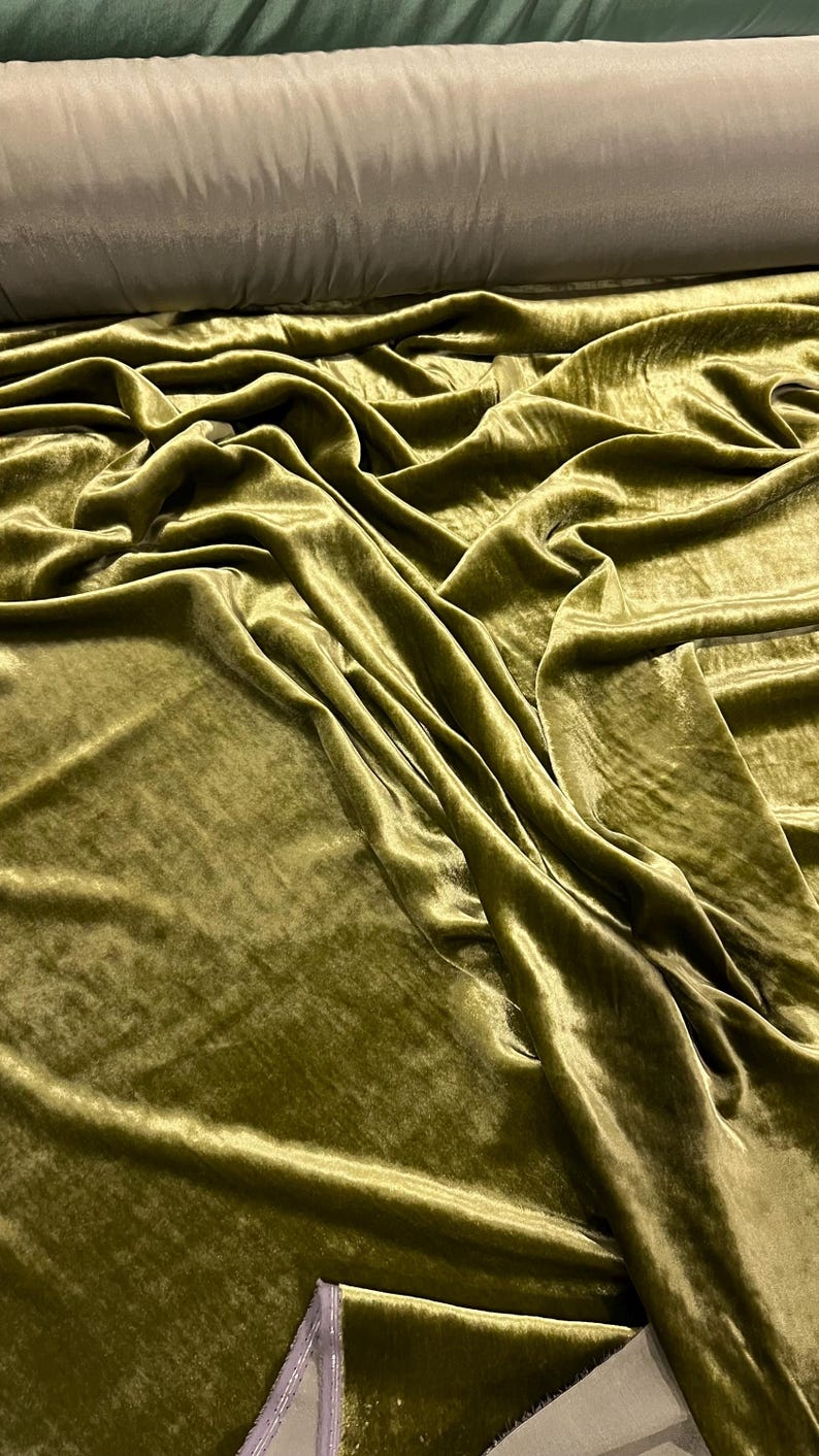 May include: A close-up of a soft, green velvet fabric. The fabric is draped and folded, showing its texture and sheen.