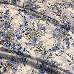 May include: White fabric with a blue floral pattern. The flowers are a variety of blue shades and are arranged in a repeating pattern. The fabric has a soft, flowing texture.
