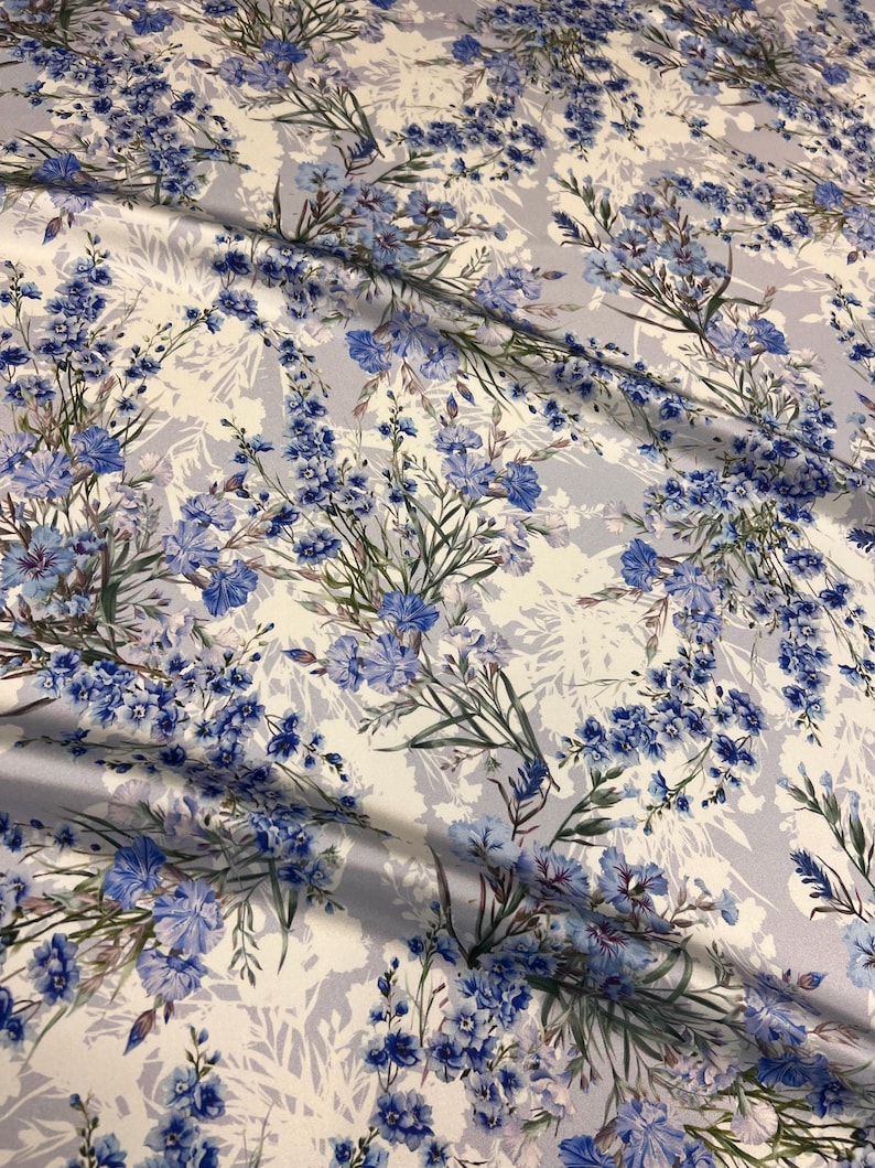 May include: White fabric with a blue floral pattern. The flowers are a variety of blue shades and are arranged in a repeating pattern. The fabric has a soft, flowing texture.