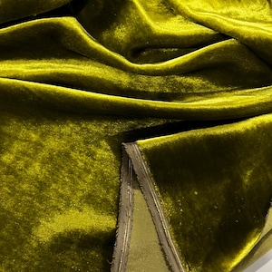 May include: A close-up of a rich, olive green velvet fabric with a subtle sheen. The fabric is folded and draped, showcasing its luxurious texture. The edges of the fabric are trimmed with a thin, gold-coloured thread.